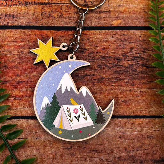 Keychain with moon design featuring mountains, tent, and star on wooden surface