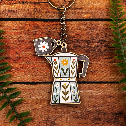 Keychain with coffee pot and mug design on a wooden surface with leaves.