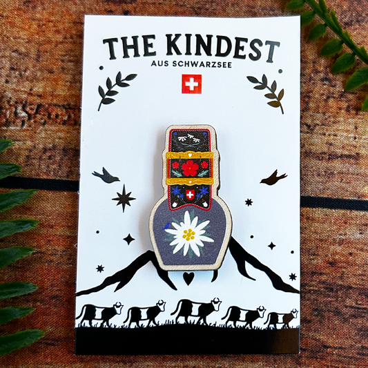 Illustrated wooden pin badge with Swiss flag design on a white card with 'The Kindest' branding, placed on a wooden surface with leaves.