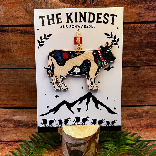 Wooden cow ornament on a card with 'The Kindest' branding against a wooden background