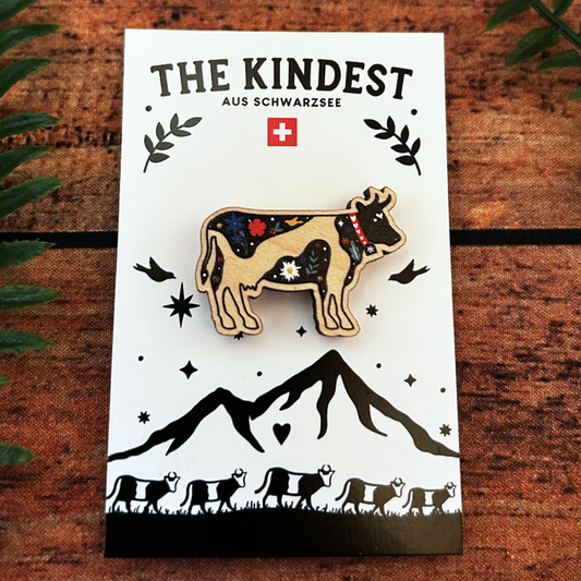 swiss cow wooden pin badge with alpine flora presented on a branded backing card.