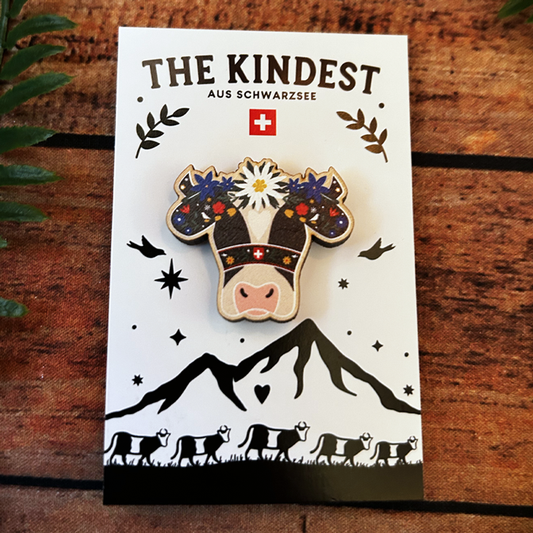wooden pin badge of a holstein cow with flowers on its head in the swiss alpabzug style presented on a backing card.