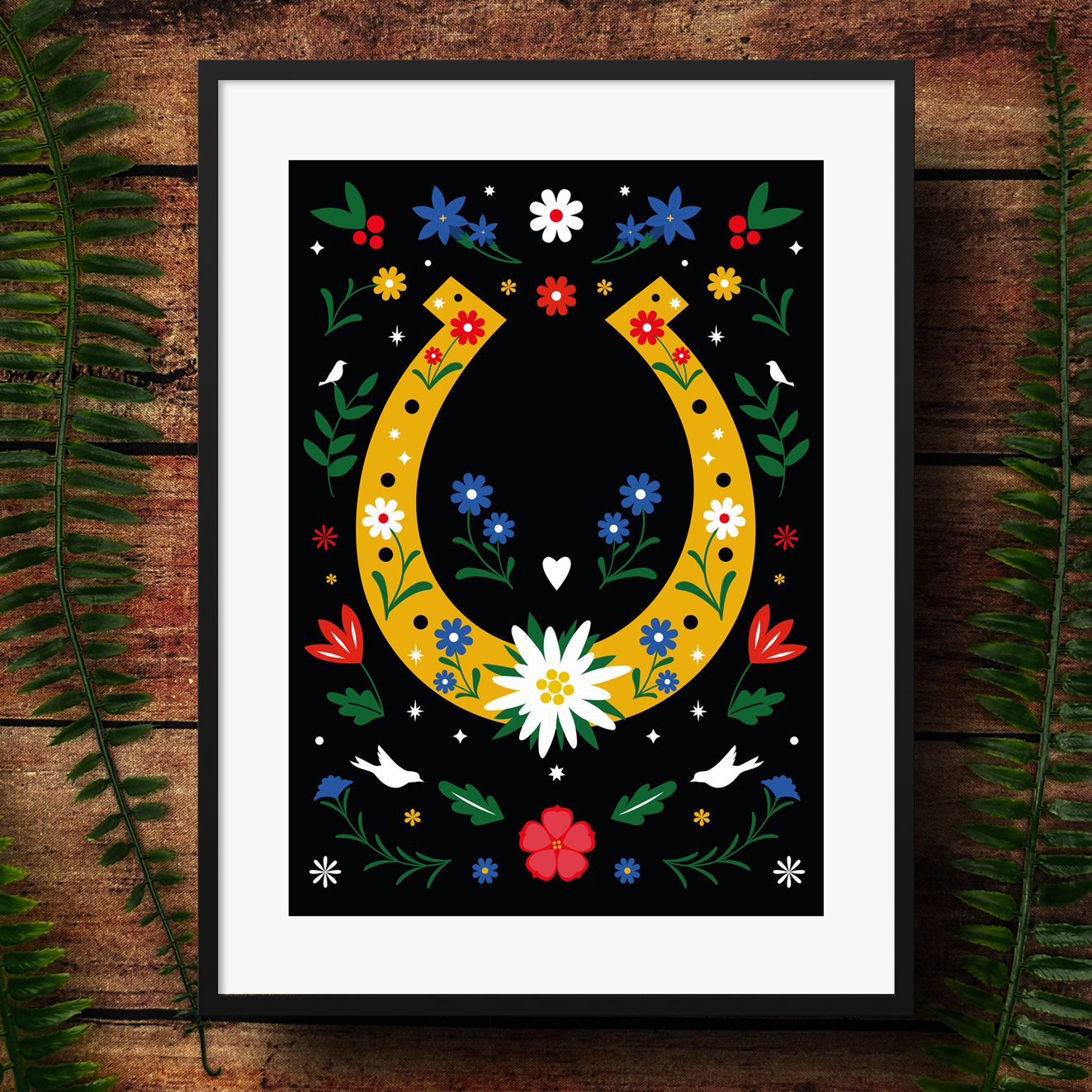 Framed artwork with a yellow horseshoe and colorful flowers on a black background, placed on a wooden surface with green leaves.