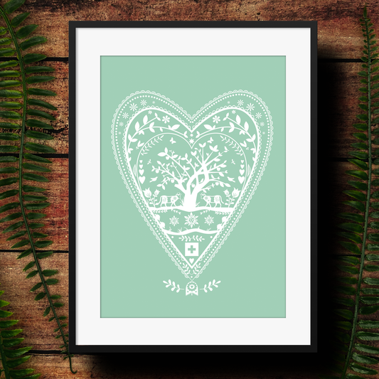 Swiss papercut style design in a heart as an art print in green and white. framed and photographed against a dark wood background.