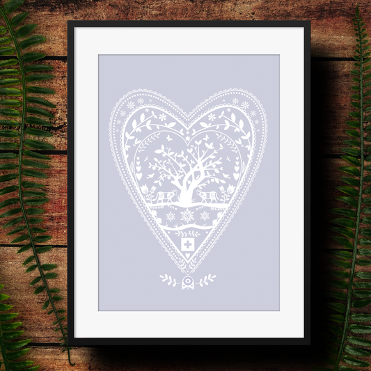 Framed artwork with a swiss papertcut white heart-shaped design on a light purple background, surrounded by green leaves on a wooden surface.