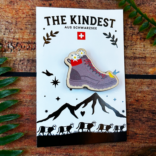 Hiking boot pin badge on a card with 'The Kindest' branding, mountains, and animals on a wooden background.