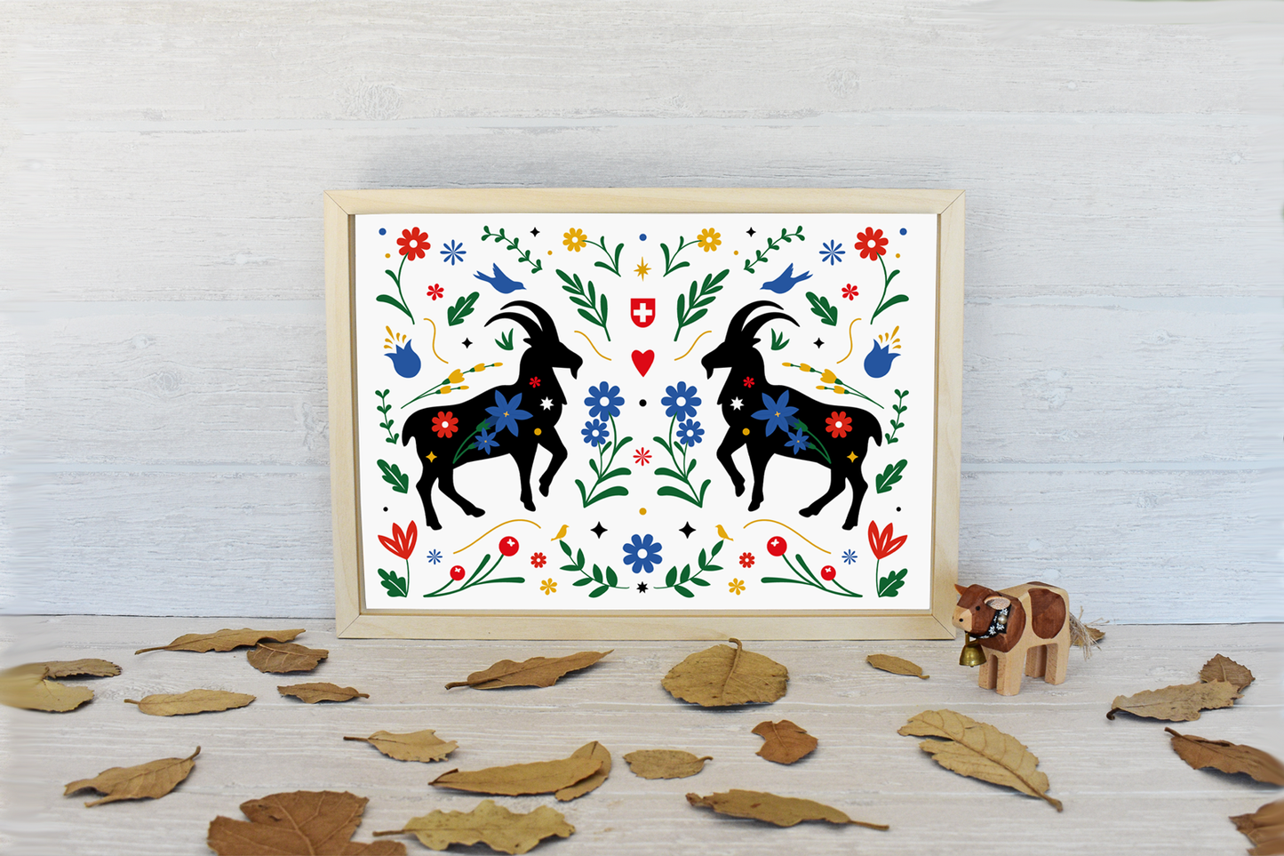 Framed artwork with two ibex and floral patterns on a white background, placed on a wooden surface with leaves.